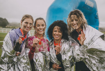 Four women are posing for the camera in foil blankets with the medals they have won from completing a charity obstacle course.