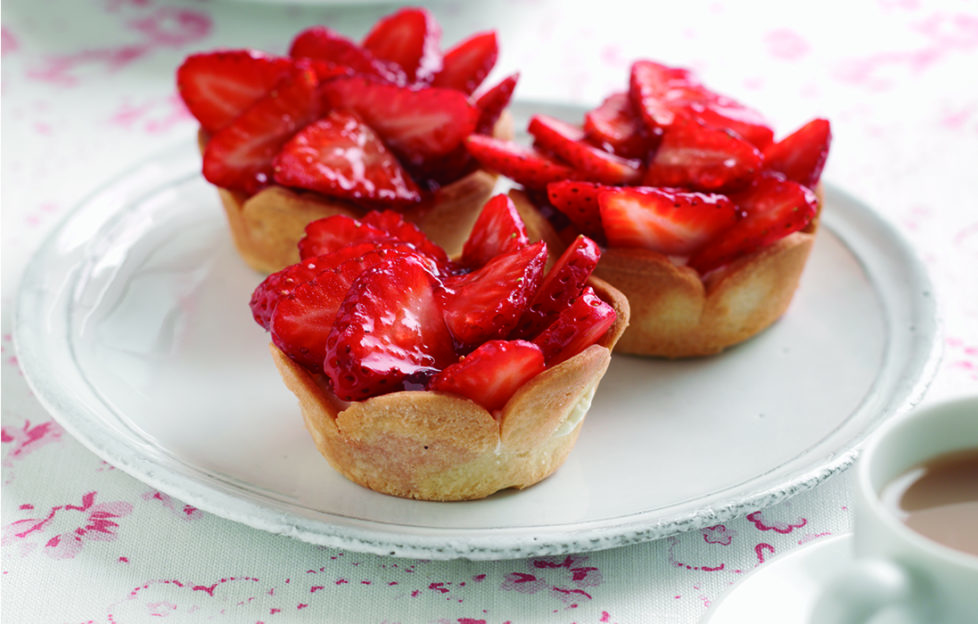 Three glazed strawberry tarts on a plate, cup of tea alongside