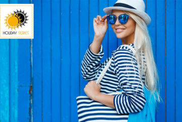 Woman in navy/white striped top and big sunhat against blue painted wooden shed, adjusting sunglasses, looking flirty