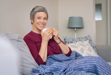 Portrait of smiling senior woman drinking coffee at home while looking at camera. Mature woman sitting on couch with warm blanket wrapped around leg. Happy lady relaxing at home with hot drink.