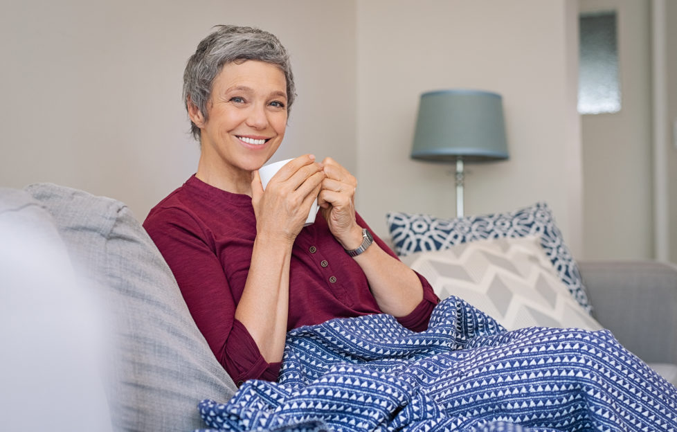 Portrait of smiling senior woman drinking coffee at home while looking at camera. Mature woman sitting on couch with warm blanket wrapped around leg. Happy lady relaxing at home with hot drink.