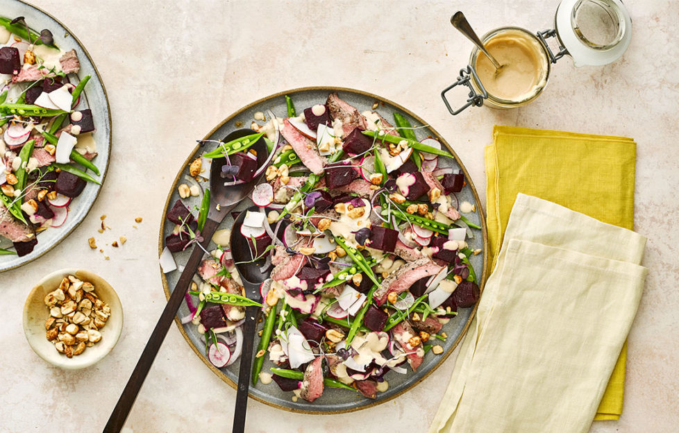 Large plate of colourful mixed salad, beetroot, sugar snaps and slices of steak, satay dressing in a dish at the side