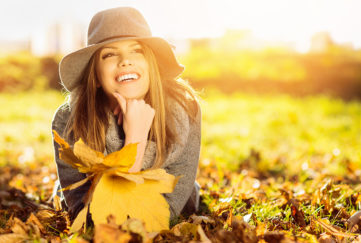 Go sober for October and feel great! Happy woman lying on stomach in autumn park, holding golden leaves, wearing floppy hat