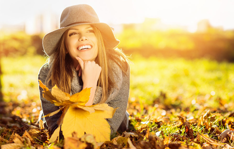 Go sober for October and feel great! Happy woman lying on stomach in autumn park, holding golden leaves, wearing floppy hat