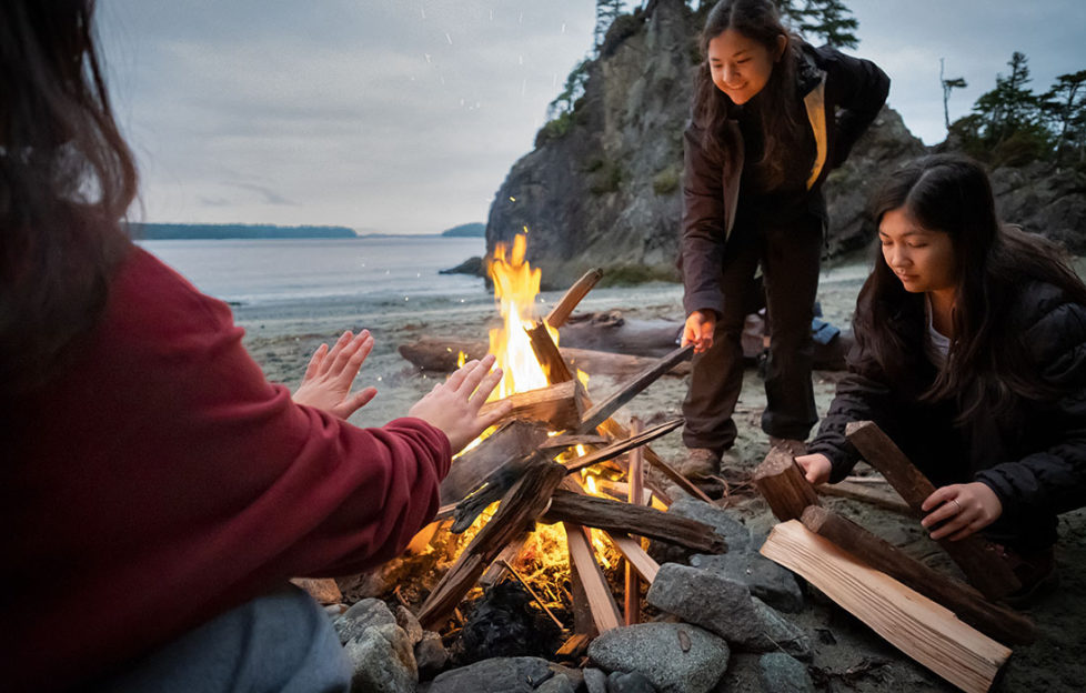 Three women around a campfire on a beach as dusk falls