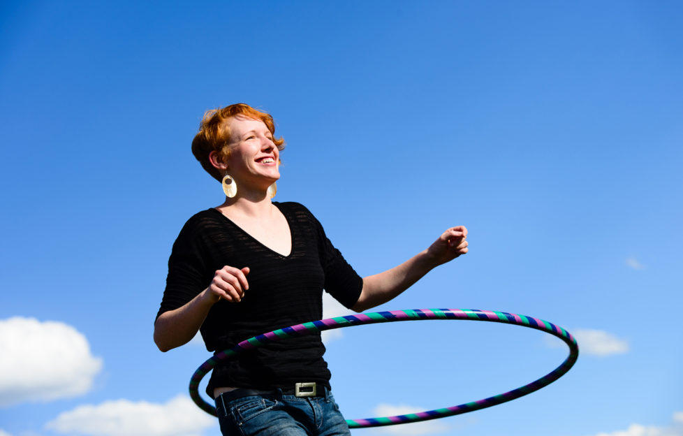 A red headed female outside playing with a hula hoop. Sky, clouds and trees in the background.