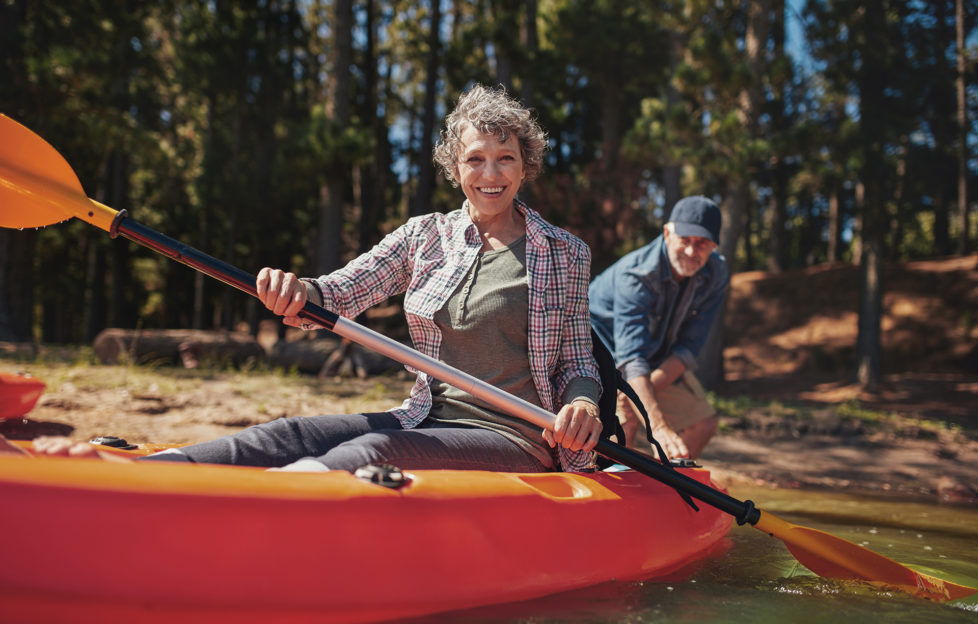 Portrait of happy senior woman in a kayak holding paddles. Woman canoeing with man in background at the lake.