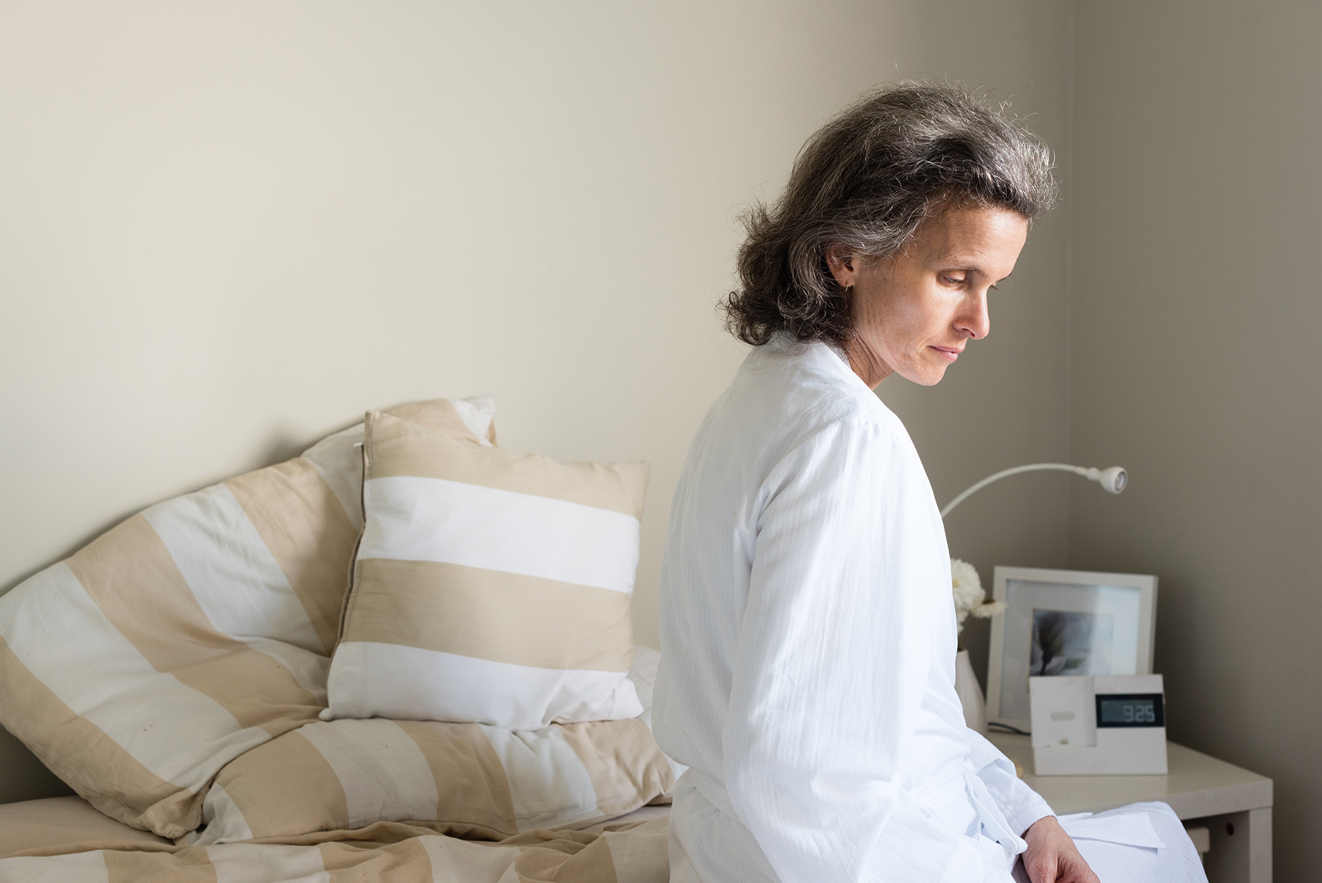 Middle aged woman in white bathrobe seated on bed looking pensive.