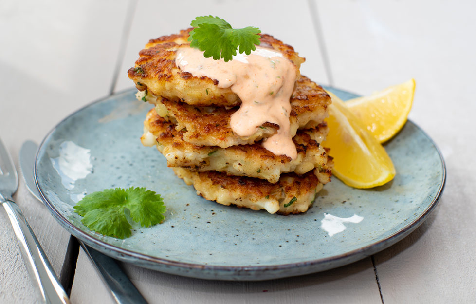 Phil Vickery recipe. Stack of 4 squid and prawn patties on a grey-blue plate, garnished with a drizzle of dip, parsley sprigs and lemon wedges