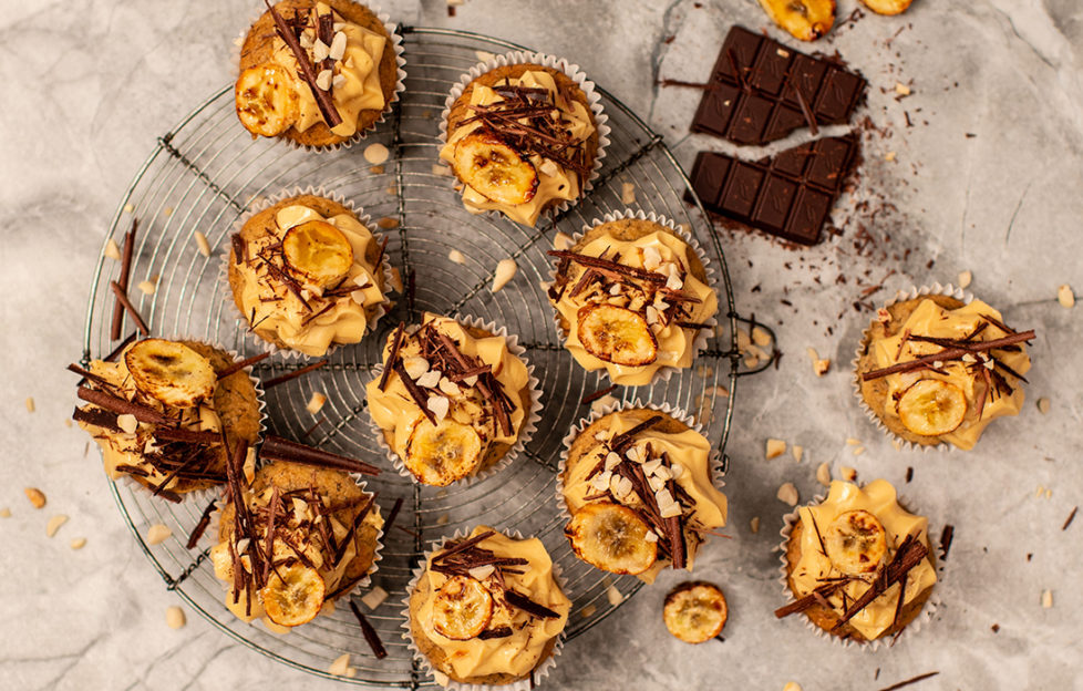 Peanut butter and banana vegan cupcakes on a cooling rack, decorated with icing, shards of dark chocolate and banana chips