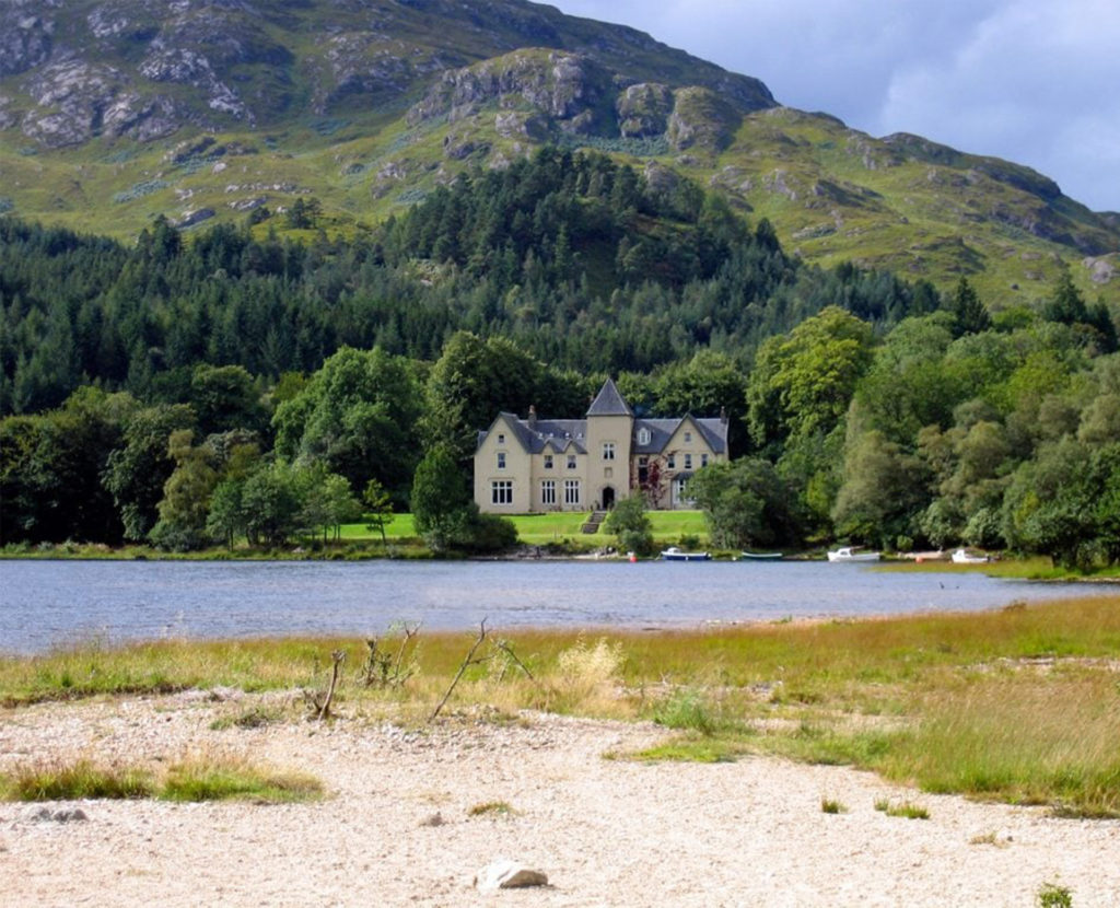 Large, grand house with tower amid trees with mountains behind, loch in foreground