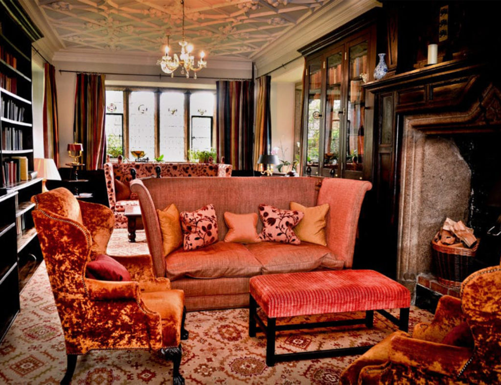 Orange silk and velvet chairs and sofa, long dark wood bookshelves, huge stone fireplace, mullioned window at end and low ornate ceiling