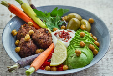 Plant power winter bowl with different coloured baby carrots, golden falafels, green rocket and avocado, red peppers, roasted chickpeas and dollop of Alpro soya yogurt swirled with red harissa paste