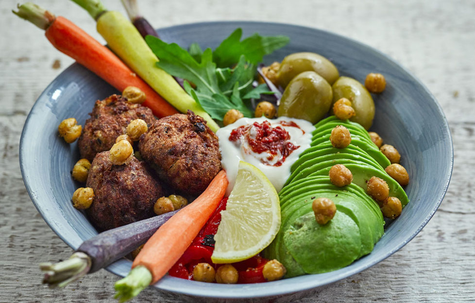 Plant power winter bowl with different coloured baby carrots, golden falafels, green rocket and avocado, red peppers, roasted chickpeas and dollop of Alpro soya yogurt swirled with red harissa paste