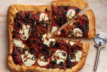 Rectangular home made pizza topped with white mozzarella slices, black ruby red tomato, sliced olives, strands of caramelised onion and capers, 2 slices cut