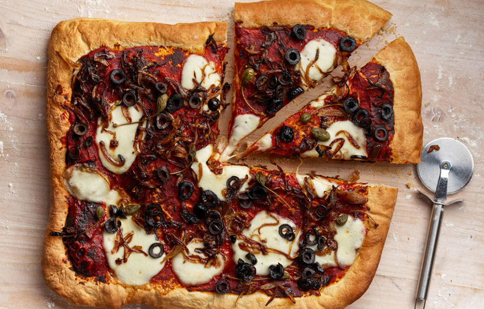 Rectangular home made pizza topped with white mozzarella slices, black ruby red tomato, sliced olives, strands of caramelised onion and capers, 2 slices cut