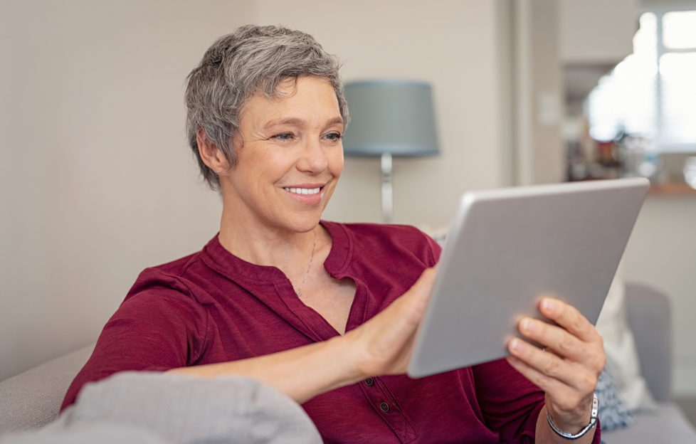 Smiling senior woman looking her digital tablet while sitting on sofa. Portrait of mature happy woman relaxing at home with digital tablet. Happy lady with gray hair browsing on laptop in living room.