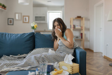 Sick Woman Covered With a Blanket Lying in Bed With High Fever and a Flu, Resting at Living Room. She is Exhausted and Suffering From Flu. Sick Woman With Runny Nose Lying in Bed. Girl Suffering From Cold Lying in Bed With Tissue Blowing Her Nose While Sitting on the Sofa