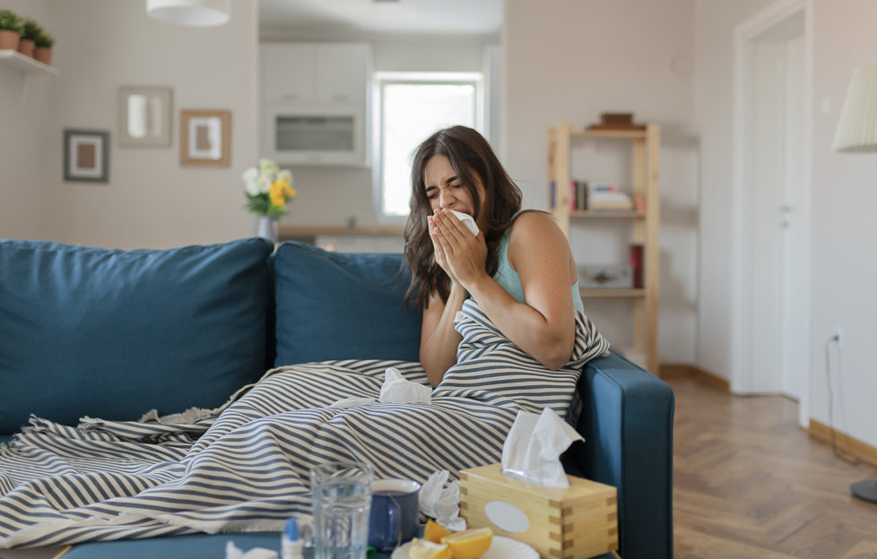 Sick Woman Covered With a Blanket Lying in Bed With High Fever and a Flu, Resting at Living Room. She is Exhausted and Suffering From Flu. Sick Woman With Runny Nose Lying in Bed. Girl Suffering From Cold Lying in Bed With Tissue Blowing Her Nose While Sitting on the Sofa