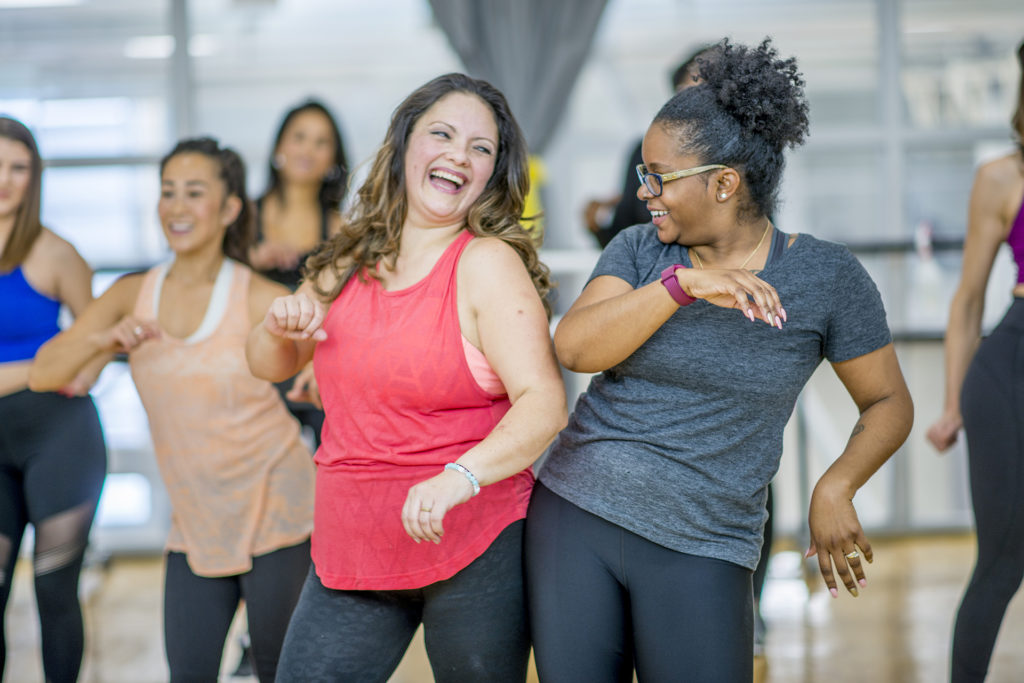 A multi-ethnic group of adult women are dancing in a fitness studio. They are wearing athletic clothes. Two women are laughing while dancing together.