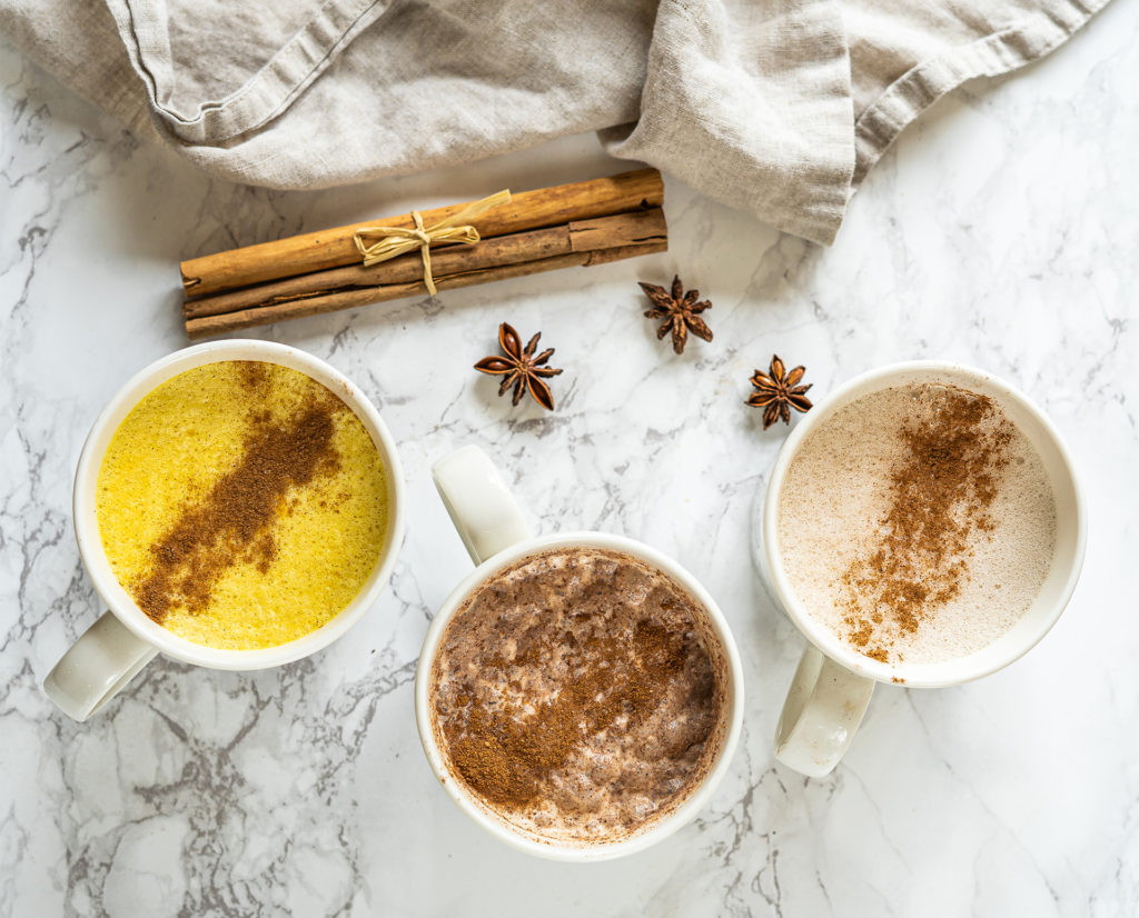 3 mugs of milk with spices sprinkled on top, cinnamon sticks and star anise to the side