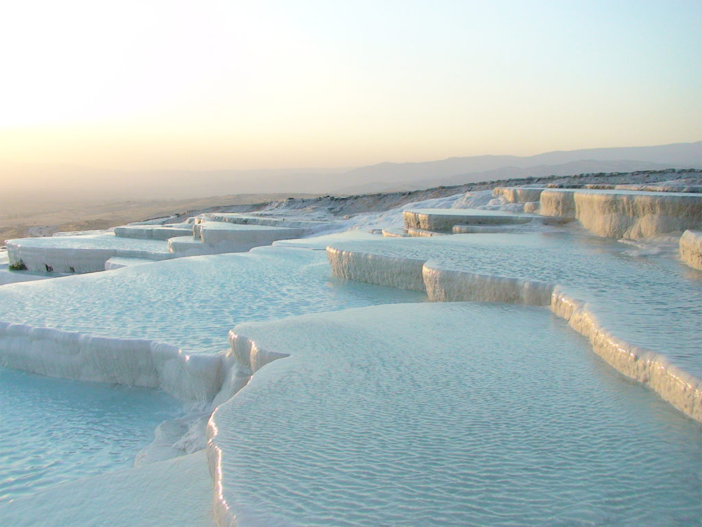 Water running gently over a series of rounded white terraces covered in mineral deposits, clear golden sky behind