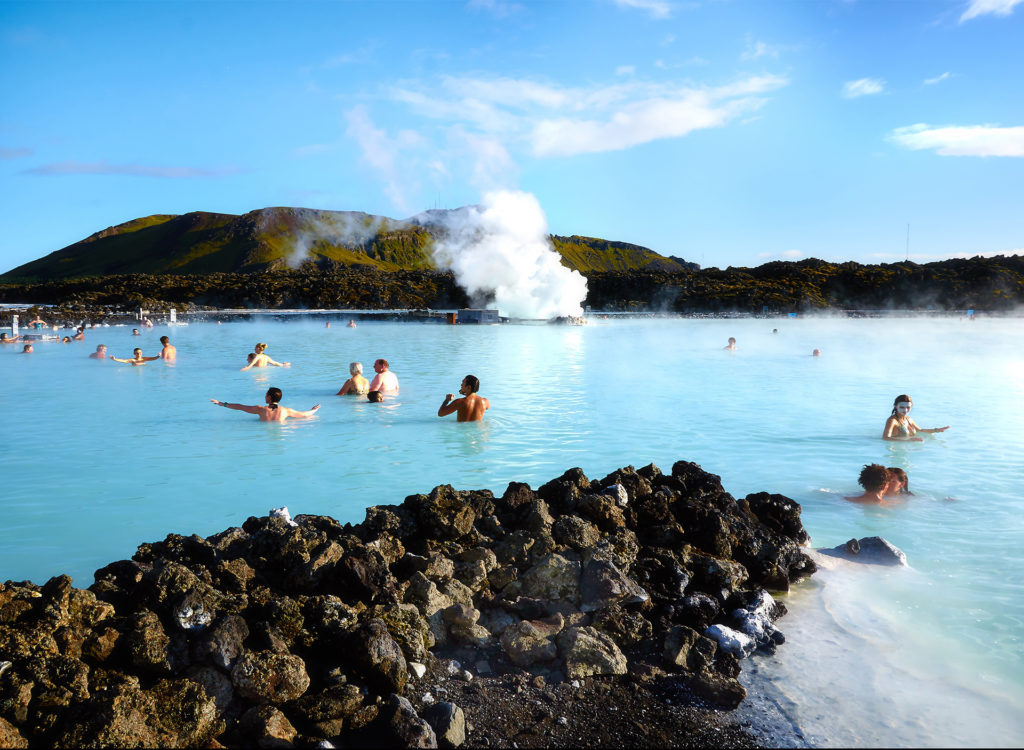 People bathing and playing in bright blue pool, rocks in foreground, hills and blue sky behind