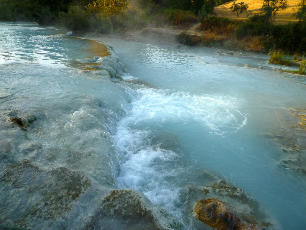 Clear turquoise water pouring over a small waterfall into a pool, Mediterranean trees and golden field beyond