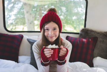A young girl wearing warm clothing holds a warm beverage and smiles.