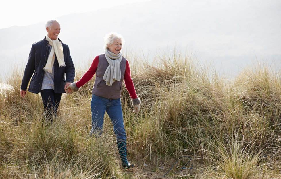 Senior Couple Walking Through Sand Dunes On Winter Beach Smiling