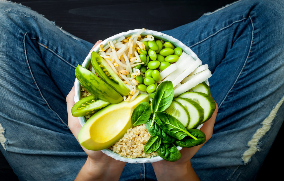 Girl in jeans holding vegan, detox green Buddha bowl with quinoa, avocado, cucumber, spinach, tomatoes, mung bean sprouts, edamame beans, daikon radish. Top view, overhead