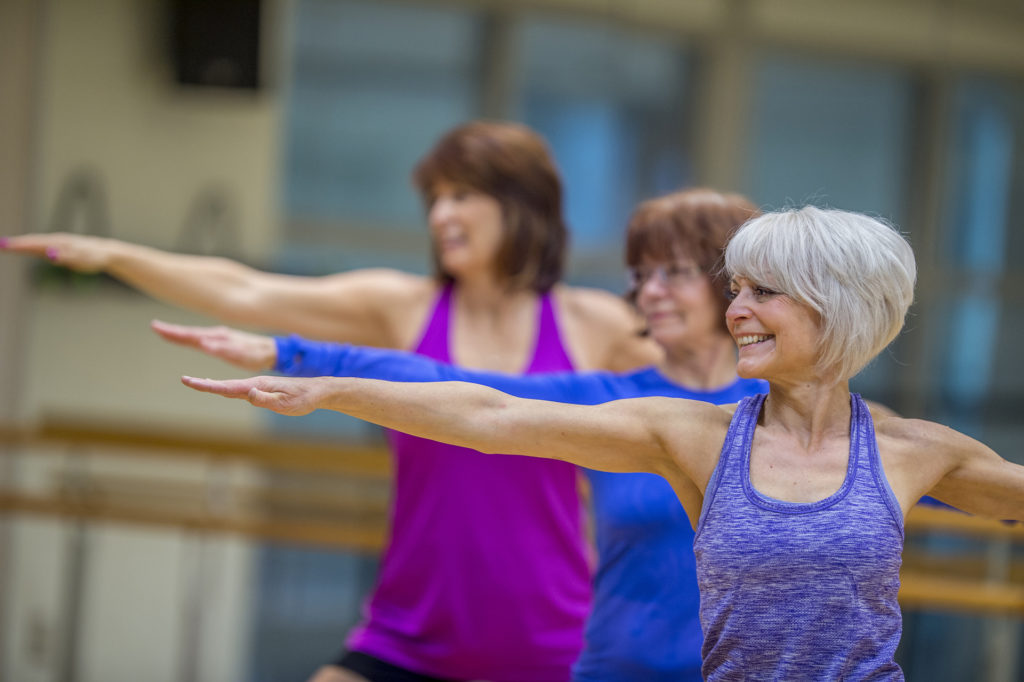 Three Caucasian senior women are indoors in a fitness center. They are wearing casual exercise clothing. They are standing and stretching their arms out while doing yoga. One woman is smiling in the foreground.