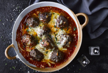 Cast iron casserole dish with baked meatballs in tomato sauce, covered in melted chees