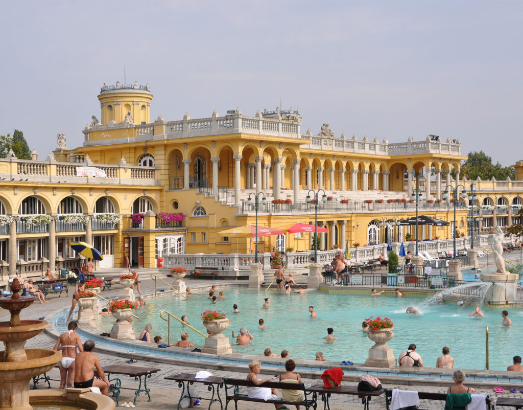 Grand European architecture, painted yellow and white, with many bathers in and around the pale turquoise water