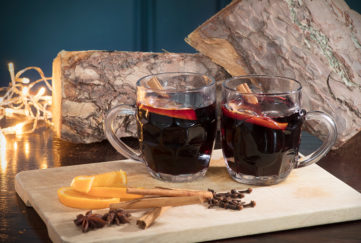 2 half pint glasses of mulled wine, dark red drink, slices of orange and spices in foreground, fairly lights and rustic log behind