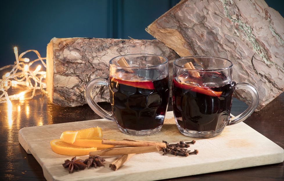 2 half pint glasses of mulled wine, dark red drink, slices of orange and spices in foreground, fairly lights and rustic log behind