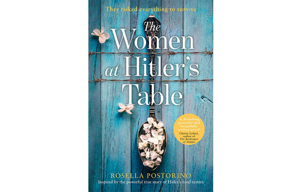 Cover of The Women At Hitler's Table, an old metal spoon lying on a blue wooden table, filled with pale pink flower petals, two strands of barbed wire running horizontally