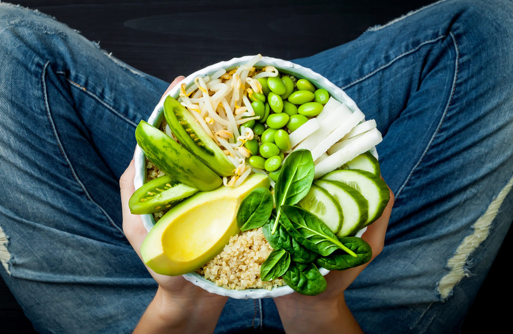 l in jeans holding vegan, detox green Buddha bowl with quinoa, avocado, cucumber, spinach, tomatoes, mung bean sprouts, edamame beans, daikon radish. Top view, overhead