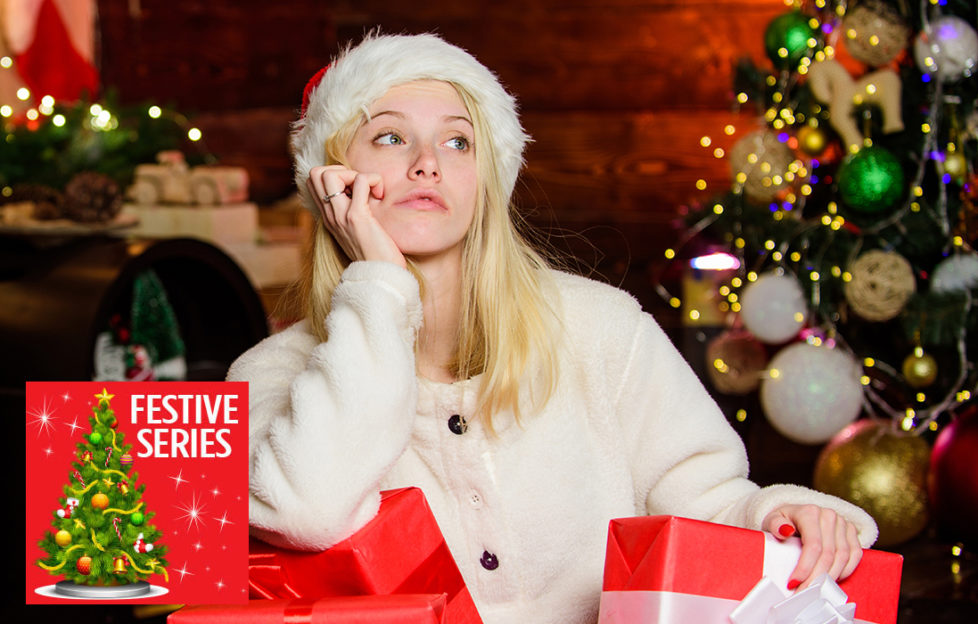 Girl in Santa hat and white jumper, surrounded by wrapped gifts, looking unhappy