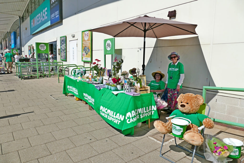  Macmillan Cancer Support. Charity stand outside superstore. Collecting money by selling china, books and cards. Three lady volunteers in uniform under white parasol.;