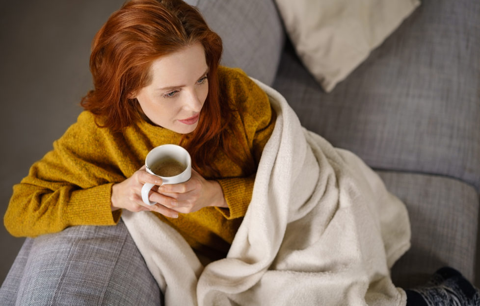 Relaxed young redhead woman enjoying a tea break sitting wrapped in a warm blanket on a comfortable couch staring thoughtfully ahead,