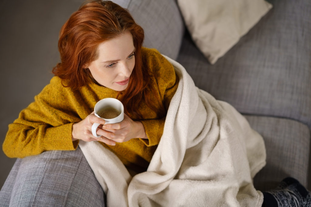 Relaxed young redhead woman enjoying a tea break sitting wrapped in a warm blanket on a comfortable couch staring thoughtfully ahead, high angle view.