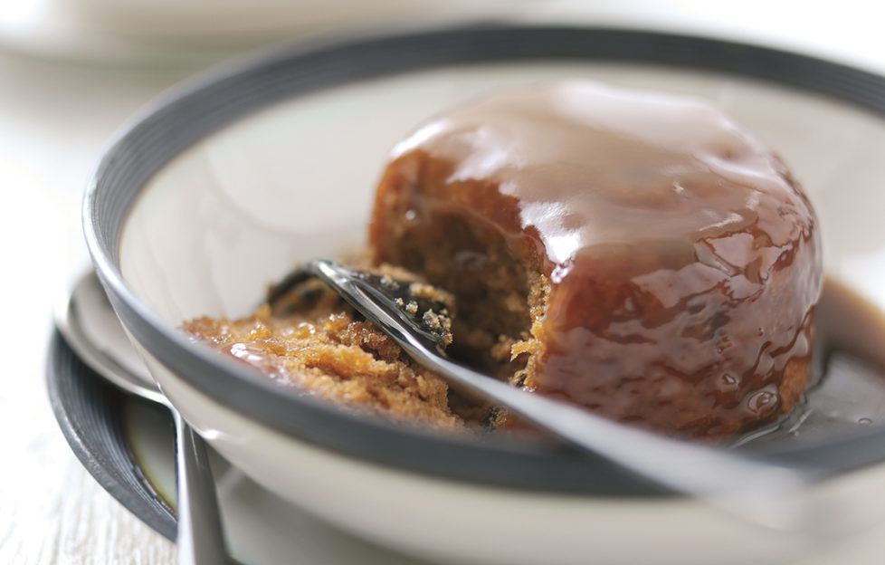 Sticky toffee pudding in a bowl, sponge coated in toffee sauce, one spoonful cut