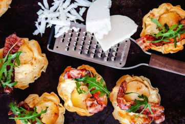 Small goat's cheese tartlets with wavy filo pastry edges, crispy bacon and a sprig of rocket, also a microplane grater with a piece of part grated cheese,