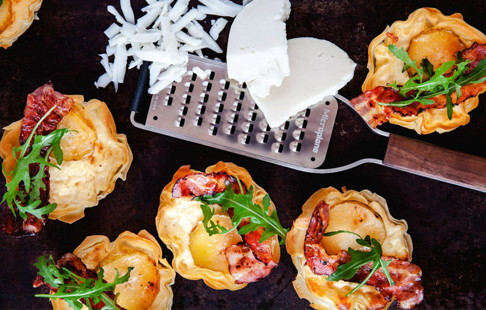 Small goat's cheese tartlets with wavy filo pastry edges, crispy bacon and a sprig of rocket, also a microplane grater with a piece of part grated cheese,