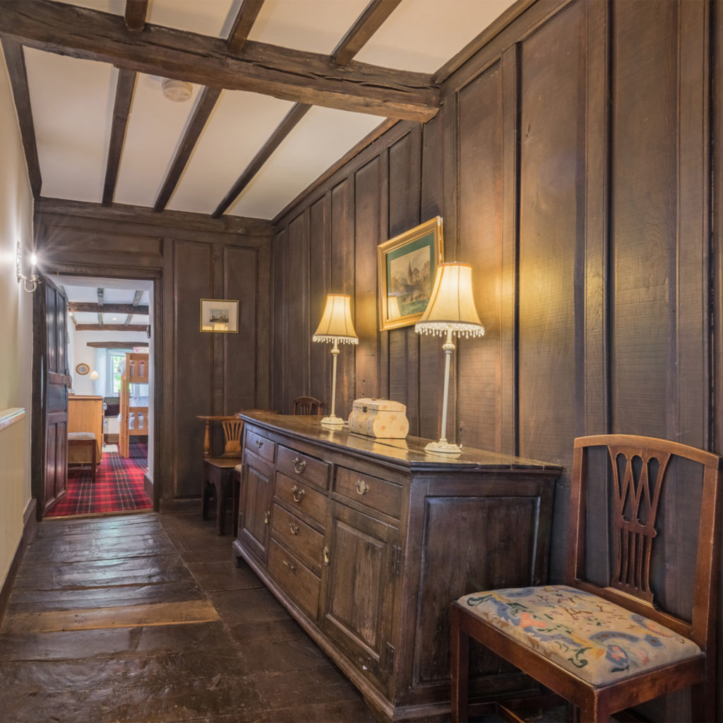 wood panelled hallway, beamed ceiling, old dresser and chairs, light bright room through doorway at the end