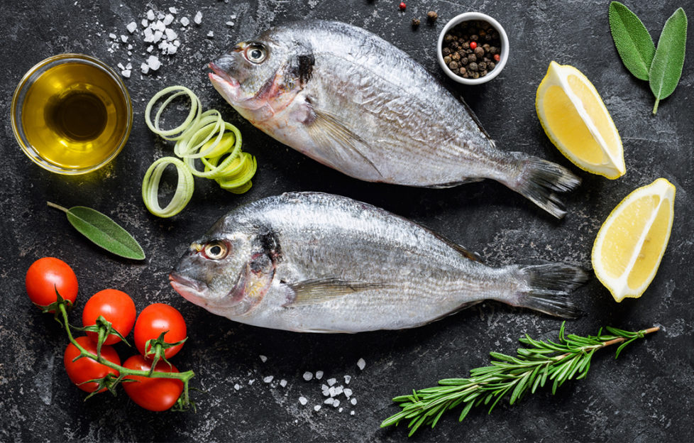 Ingredients laid out on dark granite top- 2 whole sea bream fish, tomatoes, herbs, lemon wedges