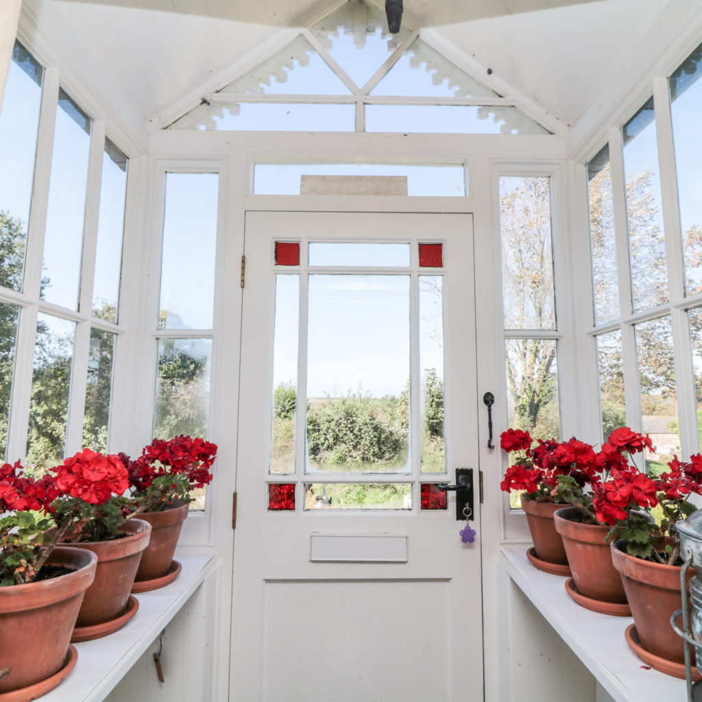 Inside glass cottage porch, pots of red geraniums on shelves, countryside views all around