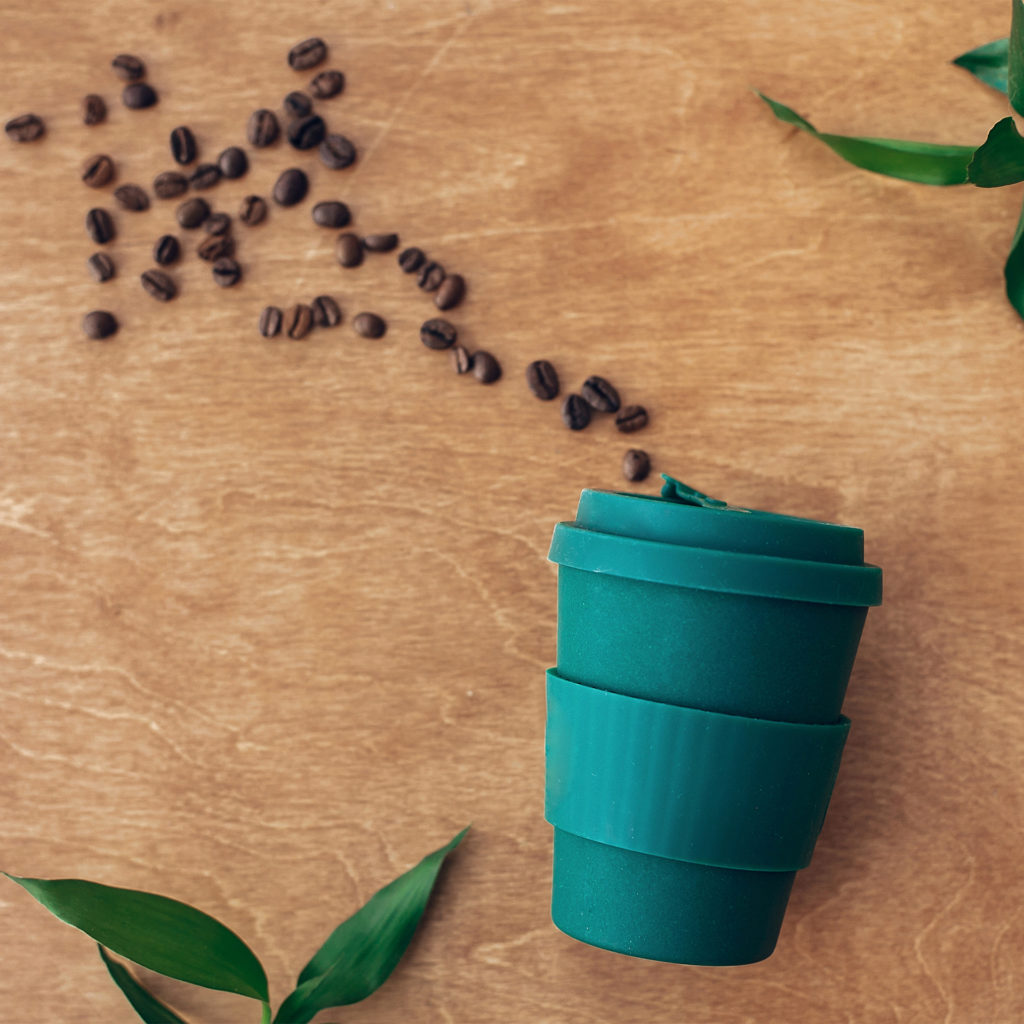 Zero waste concept, flat lay. Stylish reusable eco coffee cup on wooden background with coffee beans and green bamboo leaves. Ban single use plastic. Sustainable lifestyle. Natural bamboo cup