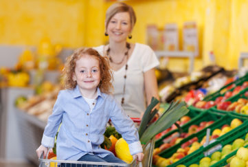 mother in a supermarket pushing a trolley filled with fresh fruit and veg with the daughter riding on the front